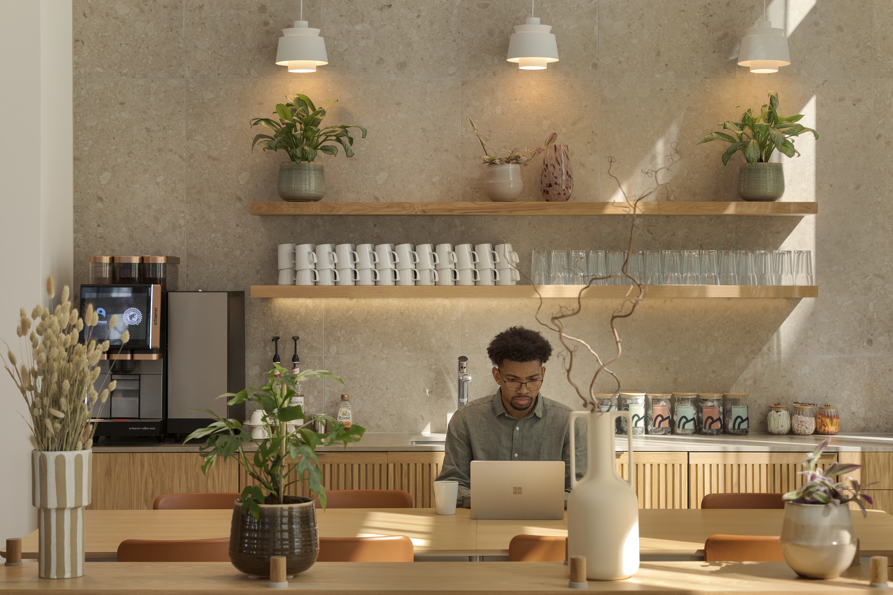 Refreshment area at MYO New Street Square featuring a coffee station, open wooden shelving with glassware and ceramics, indoor planting and an individual working from a communal table.