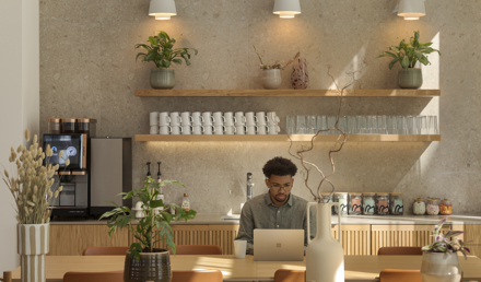 Refreshment area at MYO New Street Square featuring a coffee station, open wooden shelving with glassware and ceramics, indoor planting and an individual working from a communal table.