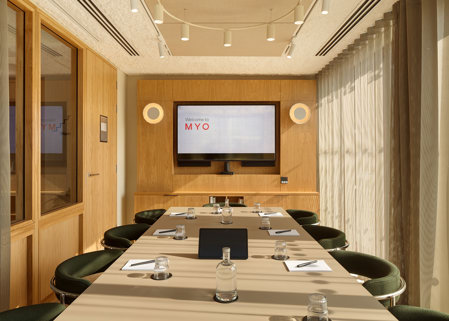 A contemporary boardroom featuring a conference table, green upholstered chairs, a presentation screen, timber wall panelling and floor-to-ceiling windows with sheer curtains.