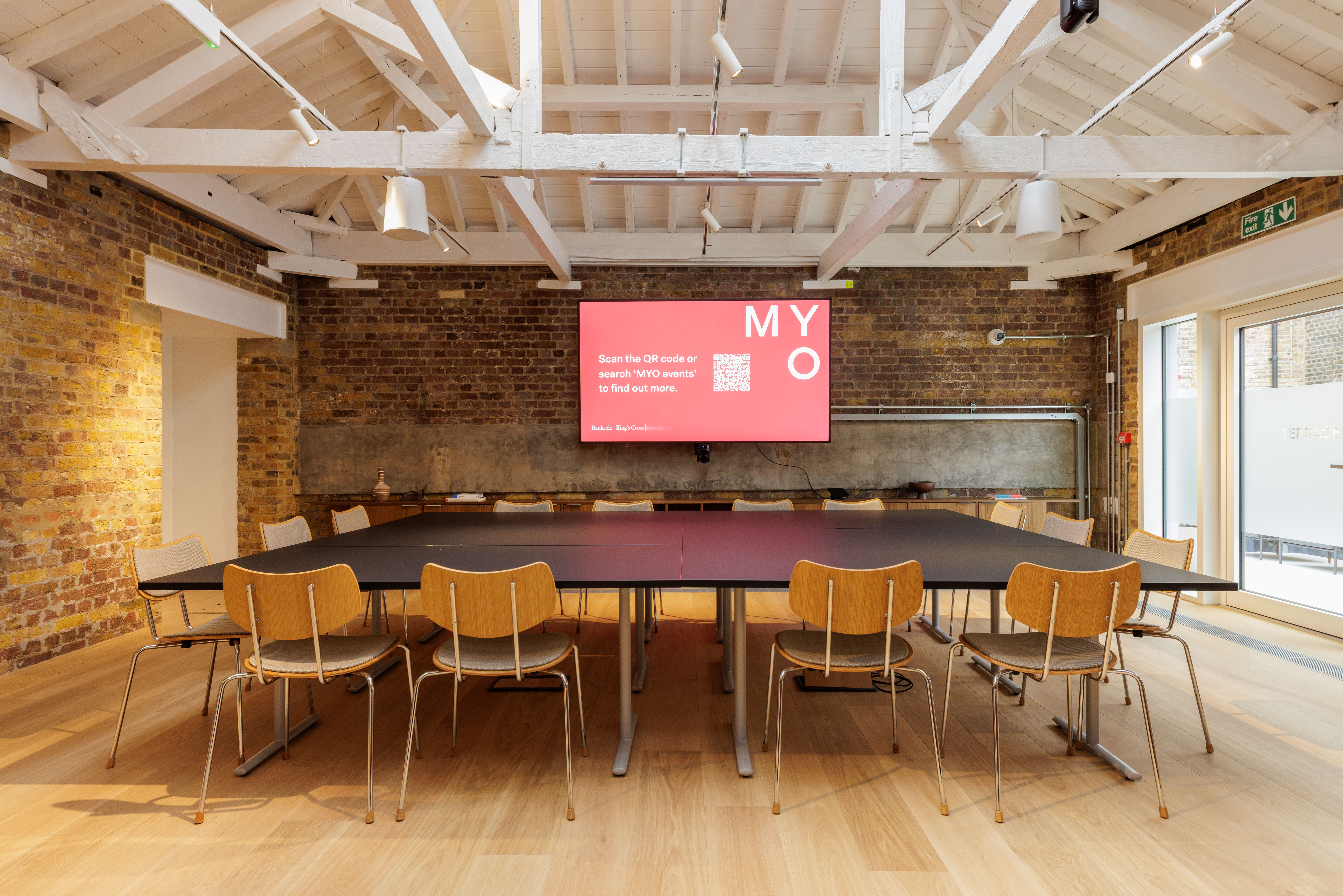 A contemporary meeting space with modular tables, wooden chairs, a large presentation screen, exposed brick walls and a vaulted ceiling with skylights.