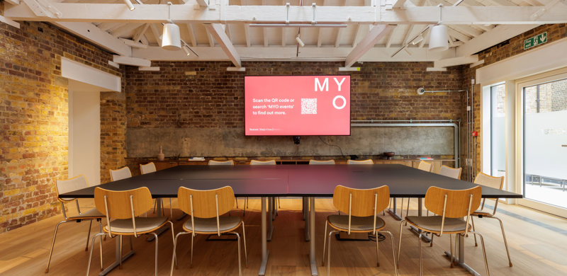 A contemporary meeting space with modular tables, wooden chairs, a large presentation screen, exposed brick walls and a vaulted ceiling with skylights.
