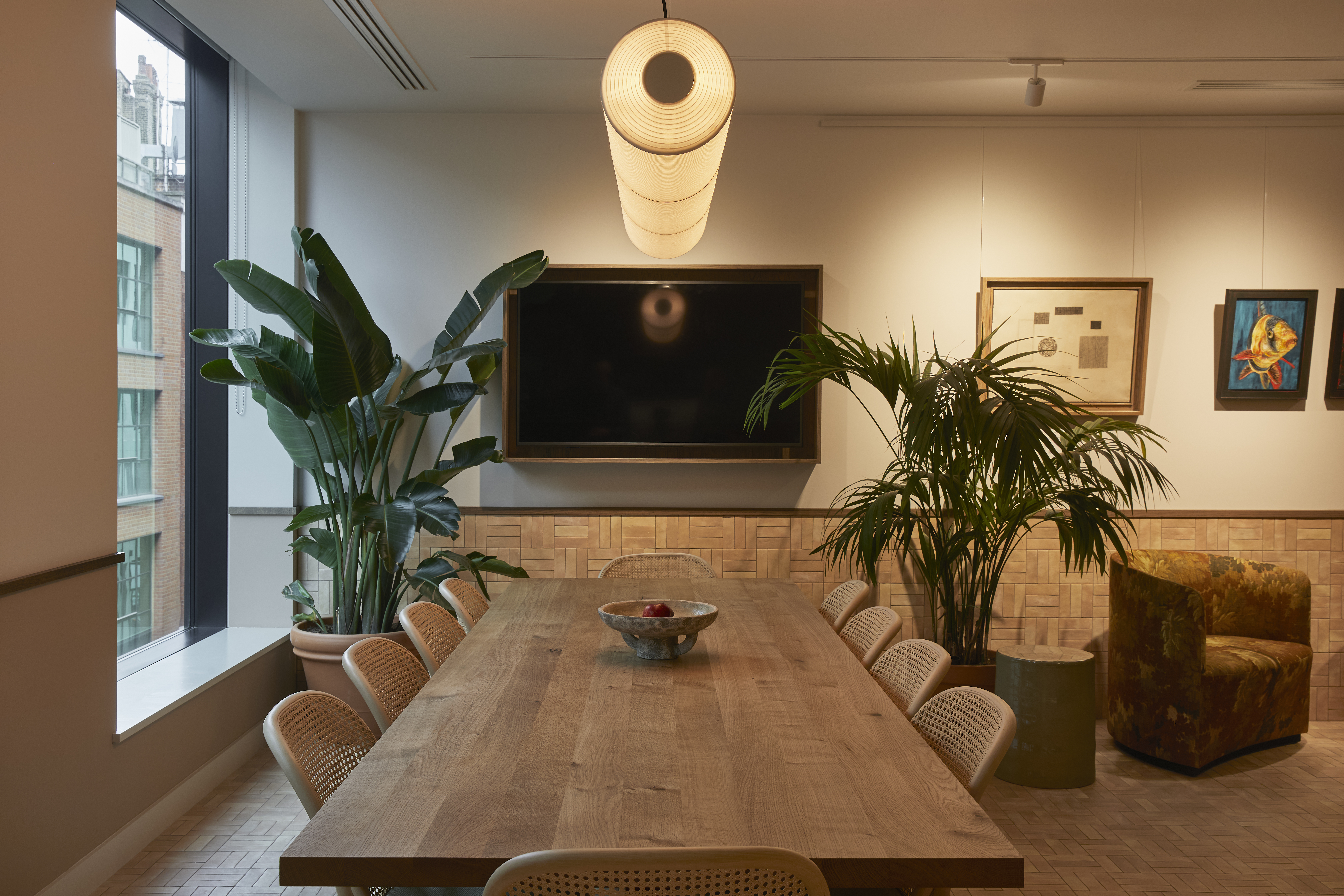 A contemporary boardroom featuring a long wooden meeting table, woven chairs, a wall-mounted display, indoor plants and modern lighting.