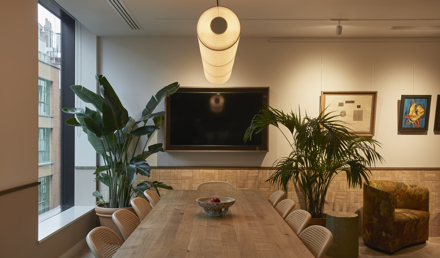 A contemporary boardroom featuring a long wooden meeting table, woven chairs, a wall-mounted display, indoor plants and modern lighting.