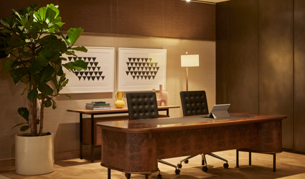 A private office featuring a wooden executive desk, two office chairs, contemporary artwork, a floor lamp and a large indoor plant.