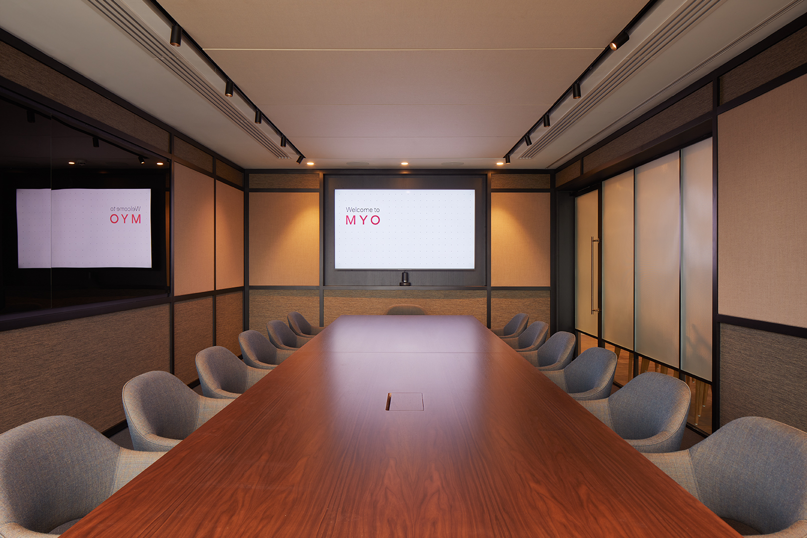 Boardroom at MYO Liverpool Street featuring a large walnut conference table, soft grey upholstered chairs, integrated presentation screen and warm ambient lighting.