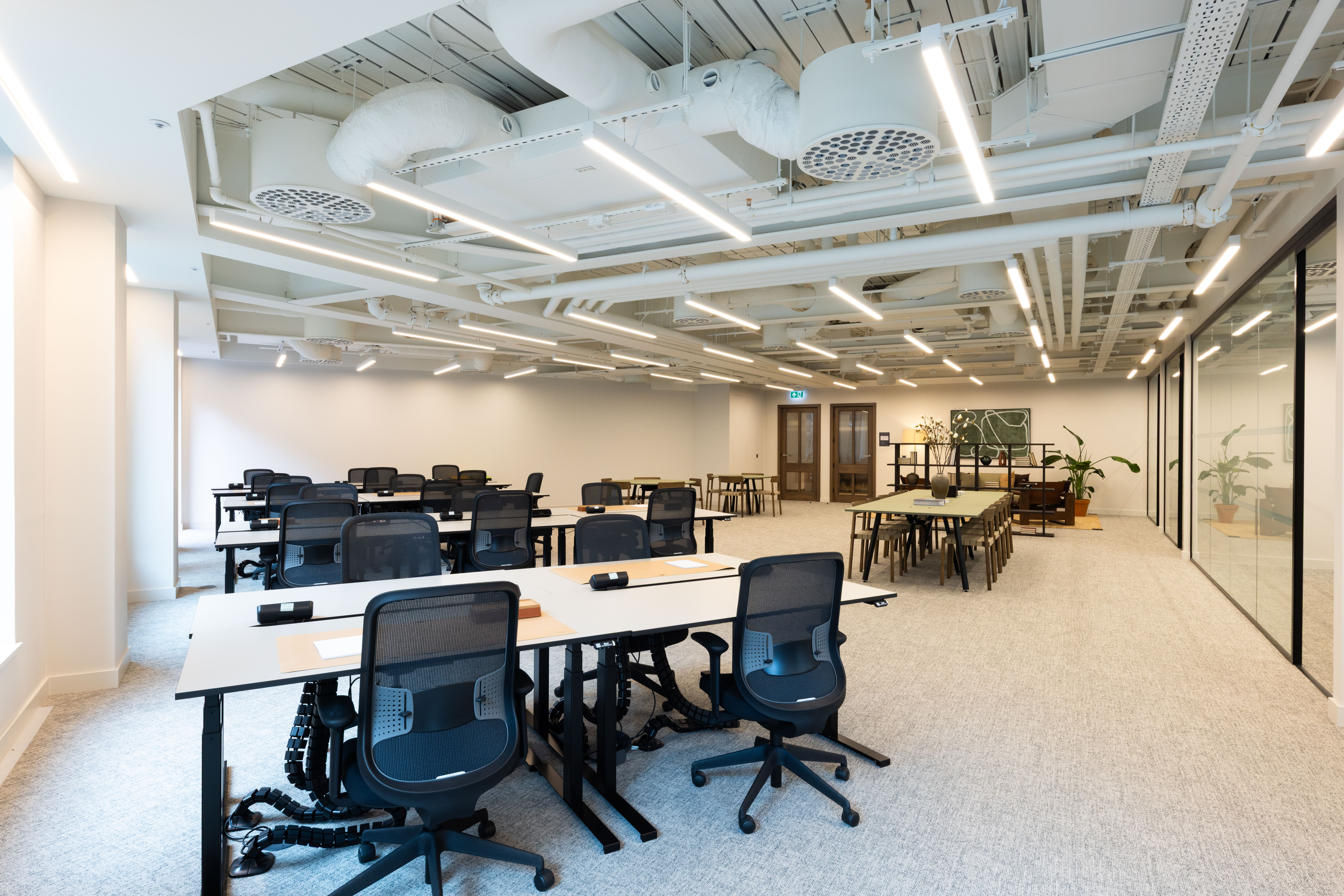 A modern open plan office featuring shared desks, ergonomic task chairs, collaborative tables and glass-fronted meeting rooms in a contemporary workspace.
