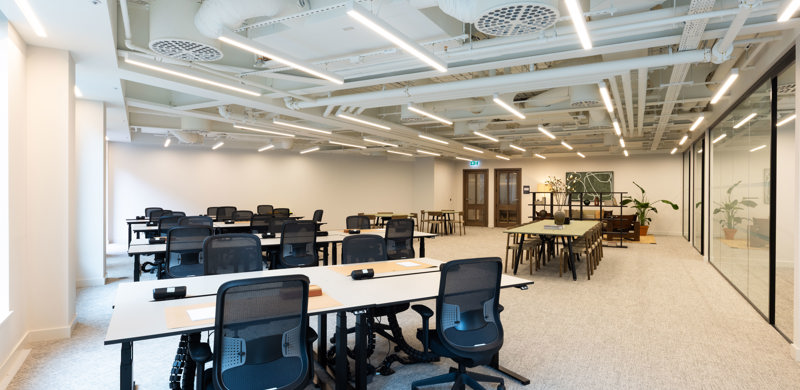 A modern open plan office featuring shared desks, ergonomic task chairs, collaborative tables and glass-fronted meeting rooms in a contemporary workspace.
