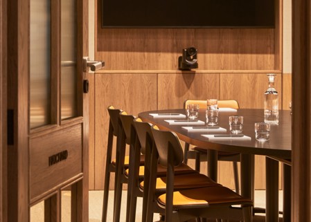 A contemporary meeting room viewed through an open doorway, featuring a round conference table, wooden chairs, video conferencing equipment and a wall-mounted presentation screen.