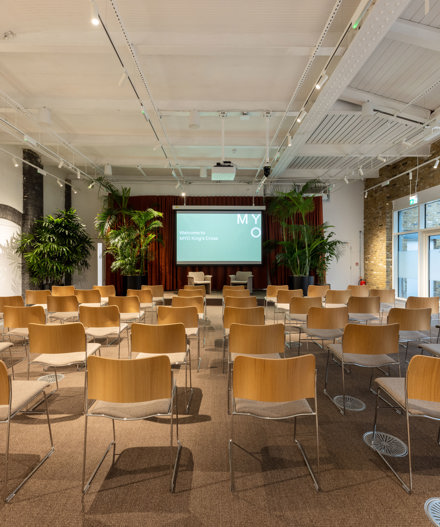 Event room with rows of wooden chairs facing a presentation screen, surrounded by indoor plants, exposed brick walls and a white ceiling with skylights.