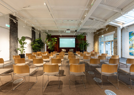 Event room with rows of wooden chairs facing a presentation screen, surrounded by indoor plants, exposed brick walls and a white ceiling with skylights.